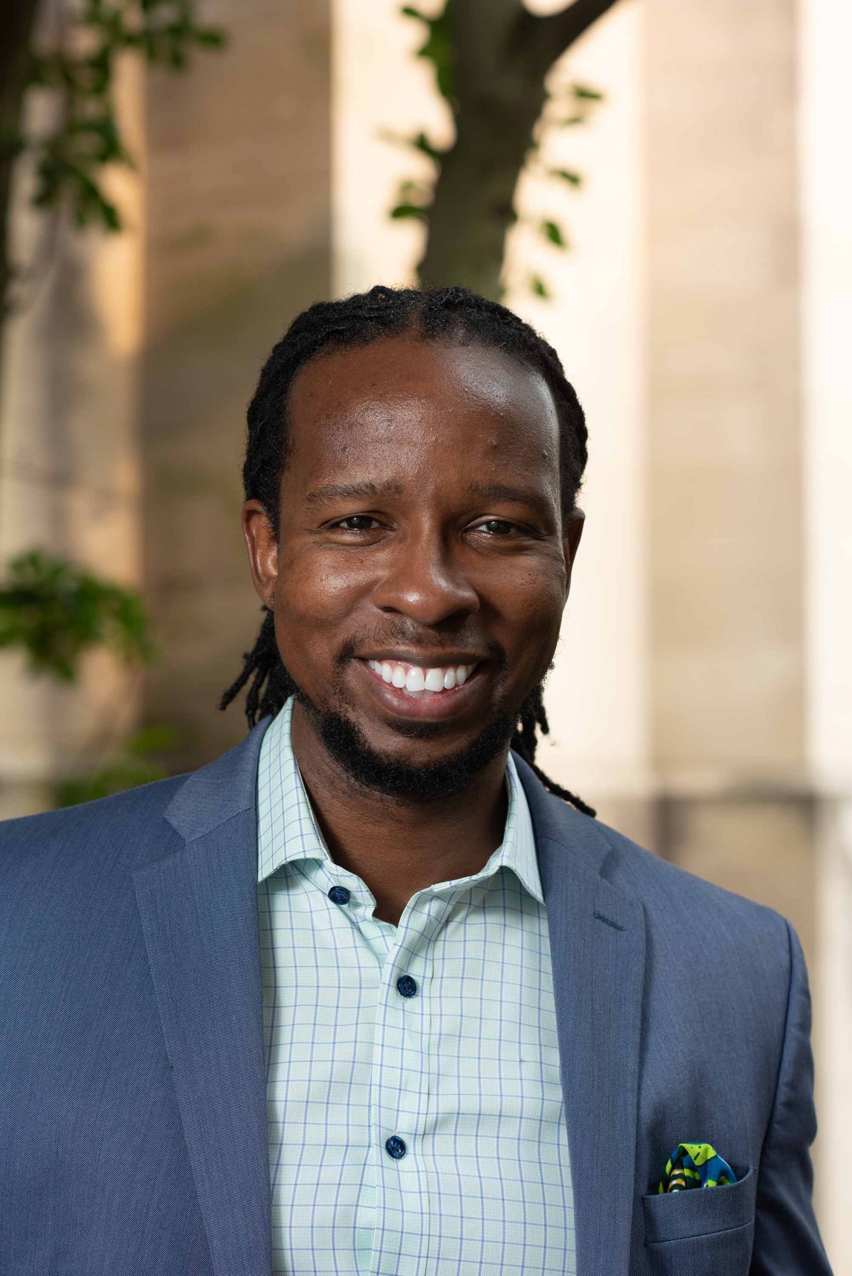 Photo of Ibram X. Kendi smiling. He has shoulder length locs that are pulled back, and he wears a blue suit, colorful pocket square, and light blue button up shirt. The portrait was taken outside, a building and a tree are blurred in the distance.