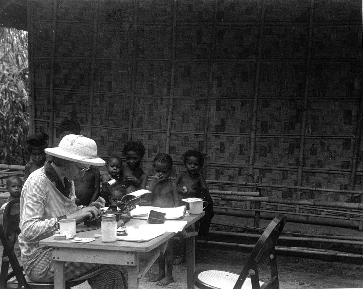 Woman wearing a white hat and glasses typing notes on a vintage typewriter as children watch.