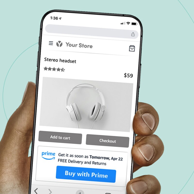 A hand holding a phone with Amazon's "Buy it with Prime" feature on the screen. Someone is buying headphones with the new feature. There is a blue text bubble on the left side of the screen that says "Buy it with Prime."