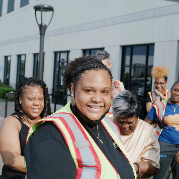 A woman wearing a safety vest smiles at the camera, as her friends and family gather behind her.