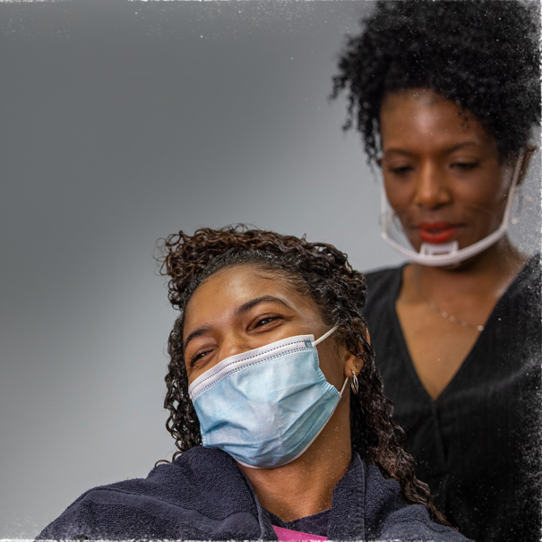 A woman wears a face mask in a stylist chair in a salon. Behind her, another woman wearing a mask is styling the first woman's hair. 