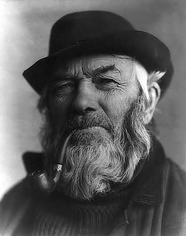 An old man looks directly at the camera with a brimmed hat and a bent-shaped pipel