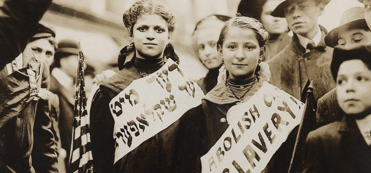 Photograph shows half-length portrait of two girls wearing banners with slogan "ABOLISH CH[ILD] SLAVERY!!" in English and Yiddish, one carrying American flag; spectators stand nearby. Probably taken during May 1, 1909 labor parade in New York City