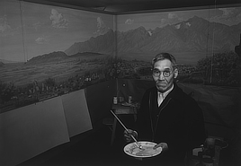 An old man wearing glasses looks directly at the camera while holding a paint brush and a plate with paint. Behind him is a panoramic scene with mountains and desert plants.