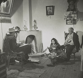 An old man sits in a chair with two young children seated on the floor in front of him  and a man and another child seated across the room. Religious symbols and portraits are on the walls, and two kerosene lamps are on the fireplace.