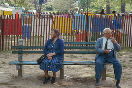 An old woman (left) and old man (right) sit on opposite ends of a bench and face away from each other.