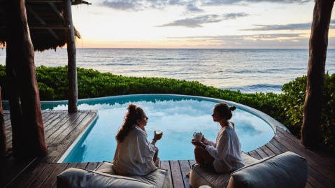 Wide shot of female friends in discussion while sitting poolside at luxury suite of tropical resort at sunrise
