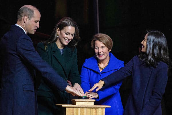 Four people stand next to one another, from left, a bald man in a navy suit, a brunette woman in a dark jacket, another woman with short hair and a bright blue jacket next to another women in a dark coat. All are smiling and have their hands on a podium pushing what looks to be a green button.