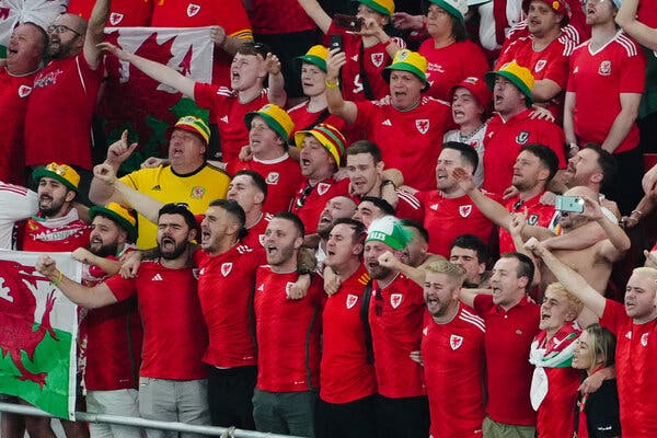 A group of jovial soccer fans stand in a stadium at the World Cup, most of them wearing red jerseys for Wales. 
