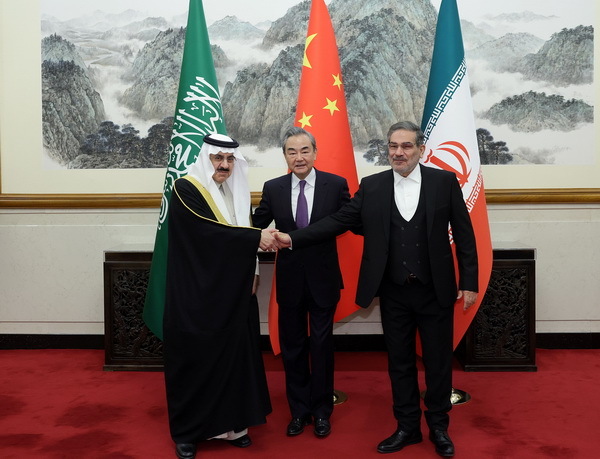 Three diplomats stand in front of their countries’ respective flags, shaking hands.