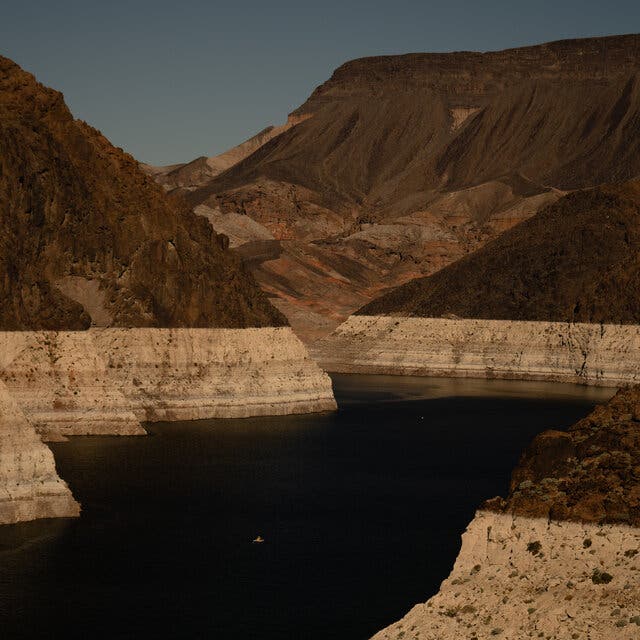 A view of  Lake Mead showing lighter colored strata of rock along the water’s surface, with greener hills above.