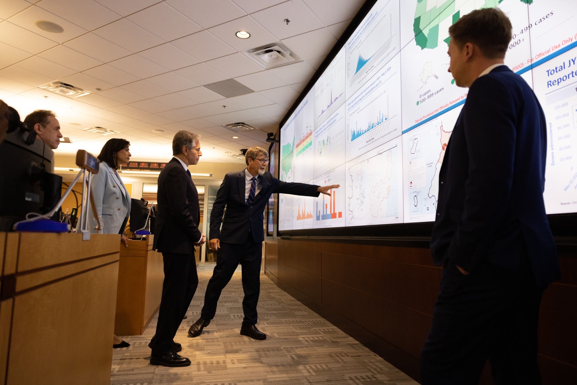 Secretary Blinken listens as a CDC official explains a graph on a screen.  The screen is part of a large wall made up of multiple screens, each displaying a different map or graph depicting health-related data in the United States.