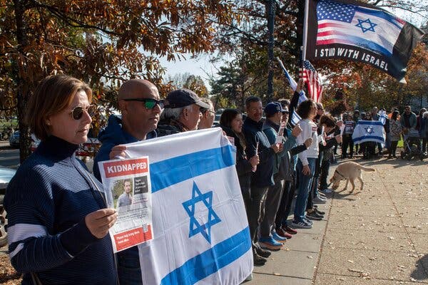 People line a sidewalk holding Israeli and American flags. A woman close to the camera holds a poster reading “Kidnapped” above the photo of a man.