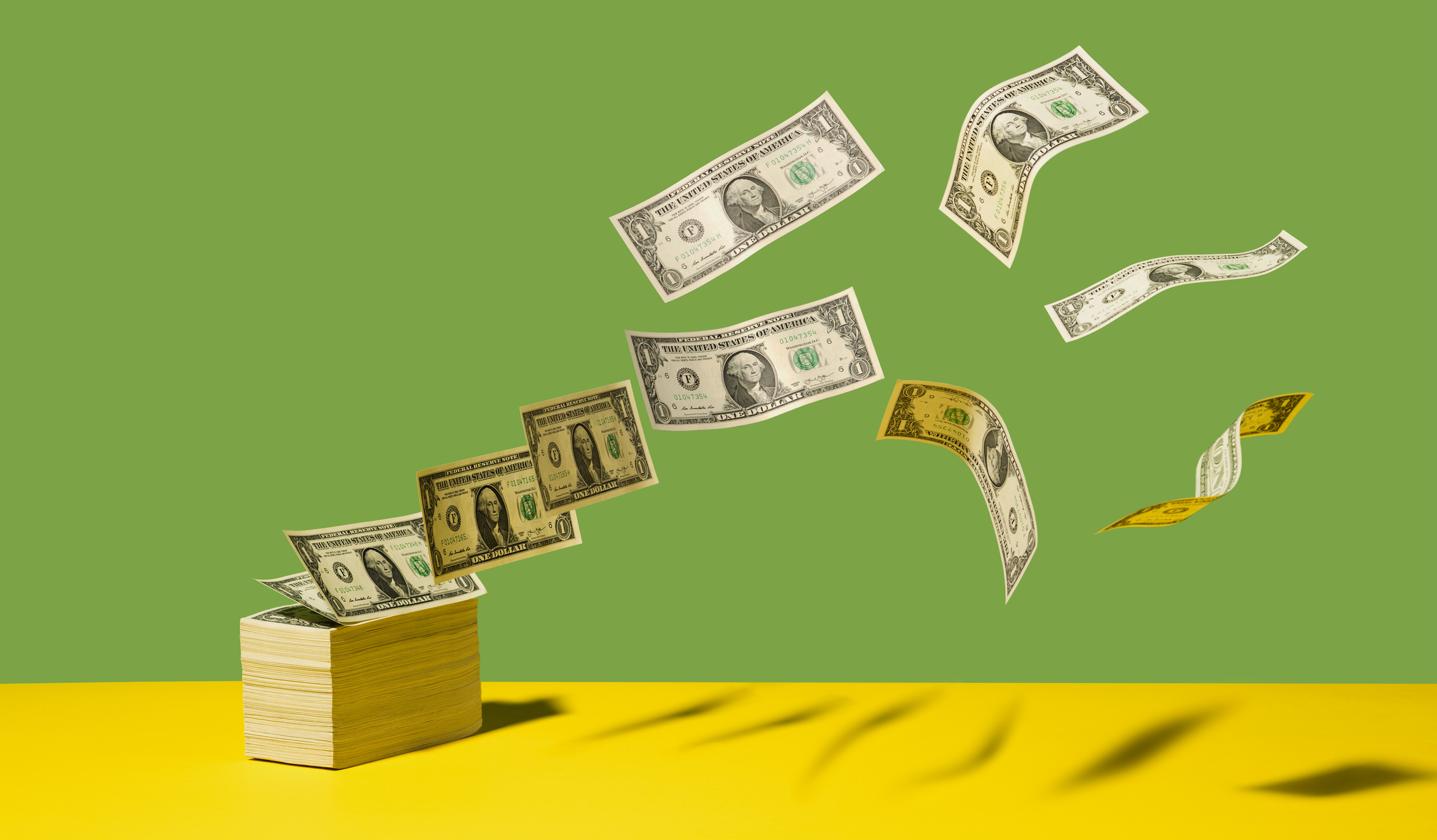 Stack of US $1 bills with bills flying away on yellow shelf, green background.