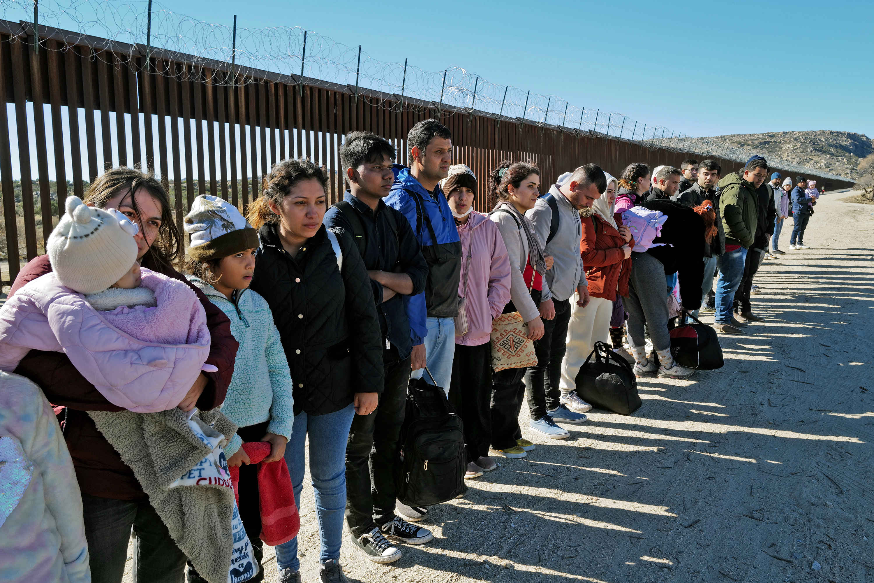 Men, women, children, and babies stand in a line before a tall fence, some with bags at their feet.