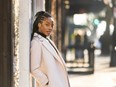 Actor Ayisha Issa, in an off-white coat, leans against a wall on a sidewalk.
