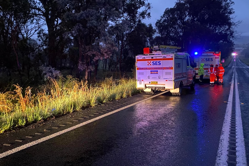 Bright red and blue lights flash on a wet roadsdie where two SES trucks are parked with emergency workers on the road