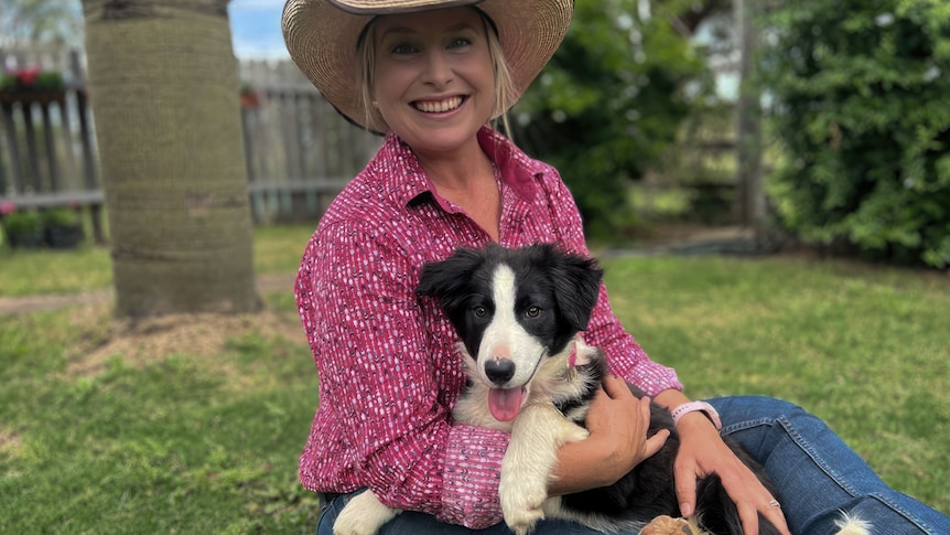 Woman in pink check shirt and broad-brimmed hat cuddles puppy and sits on the grass