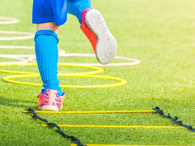 A child soccer player's feet with soccer boots on training on agility speed ladder laid out on grass.