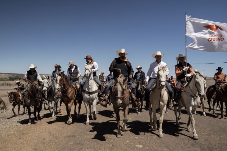 A line of political candidates and their supporters ride horses in the north Mexican desert.