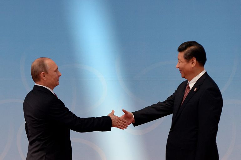 Putin and Xi greeting each other at a conference in Shanghai in 2014. They are in front of a pale blue backdrop and shaking hands.