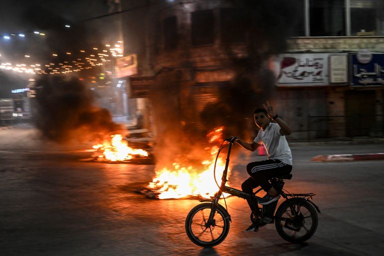 A Palestinian youth flashes the V for victory sign as he rides his bicycle next to burning tyres during an Israeli raid on the occupied West Bank city of Jenin on May 21, 2024. - Palestinian health officials said seven Palestinians were killed on May 21, in an Israeli raid on the West Bank city of Jenin, where an AFP correspondent reported masked gunmen later exchanged fire with Israeli forces. (Photo by RONALDO SCHEMIDT / AFP)
