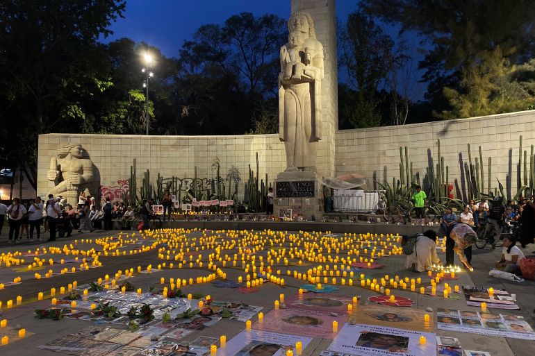 A photo of the Mother's Monument in Mexico City, an obelisk with the statue of a mother holding a child at its base. In front of the monument, the floor is filled with photographs and candles, as families seek justice for their missing loved ones.