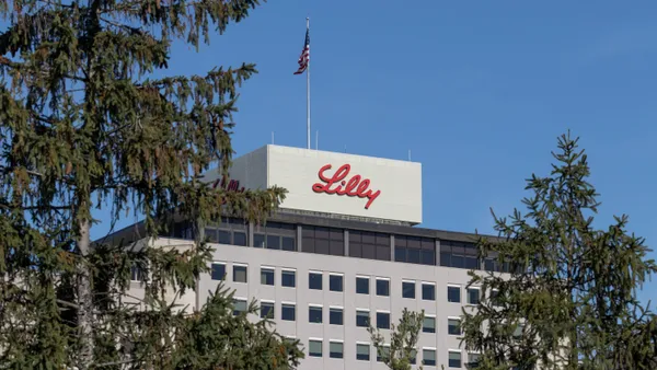 A Lilly sign is seen on the side of a building viewed through pine trees