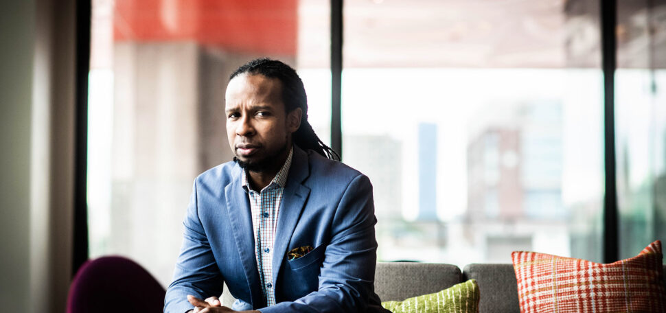 Photo of Ibram X. Kendi, a Black man with with long dreadlocks sits in a blue suit looking out a window.
