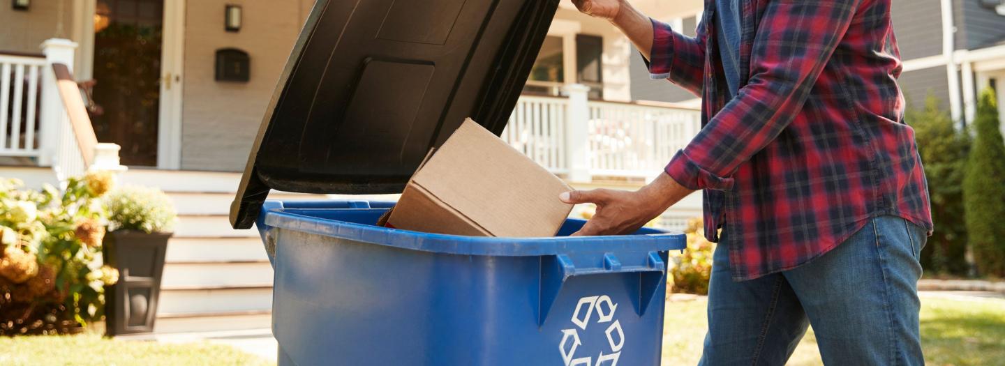 A man puts a cardboard box into a blue recycling bin at the curb