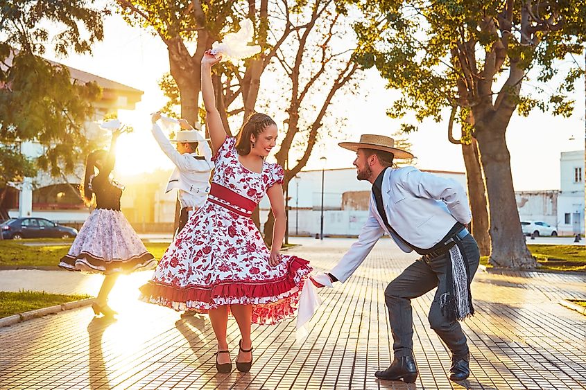 In La Serena, Chile, young adults in huaso costumes dance cueca at sunset in the city square, celebrating a national holiday. The scene is vibrant with joy and romance, surrounded by nature and city buildings.