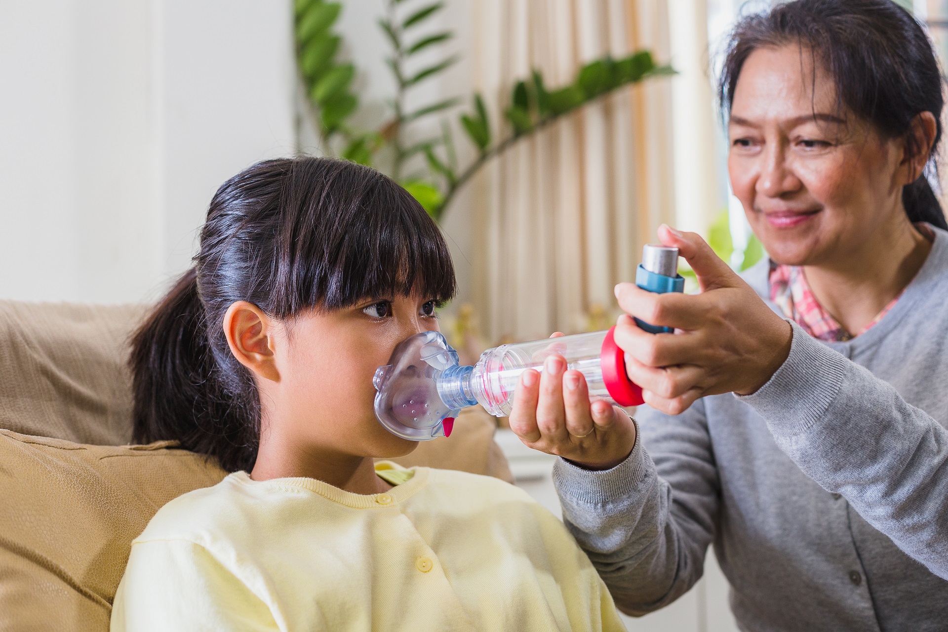 Asian mother assisting her daughter to get help with asthma inhaler spacer while sitting on sofa in living room at home