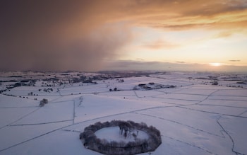 Snow showers drift in as dawn breaks over a neolithic burial mound and ring of trees in the Derbyshire Peak District near Parwich kknown as Minninglow