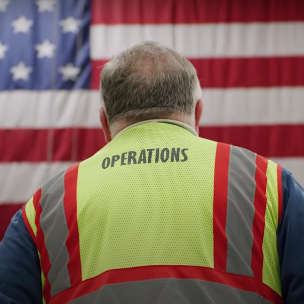 An image of an Amazon employee in a yellow work vest standing in front of an American flag.