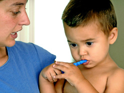 a toddler reluctantly brushing his teeth while his mum holds and encourages him
