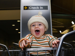 excited baby in a luggage trolley in front of a check-in sign at an airport