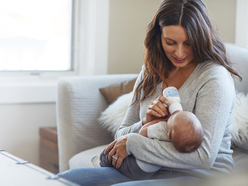 happy woman sitting on grey couch and bottle feeding a baby