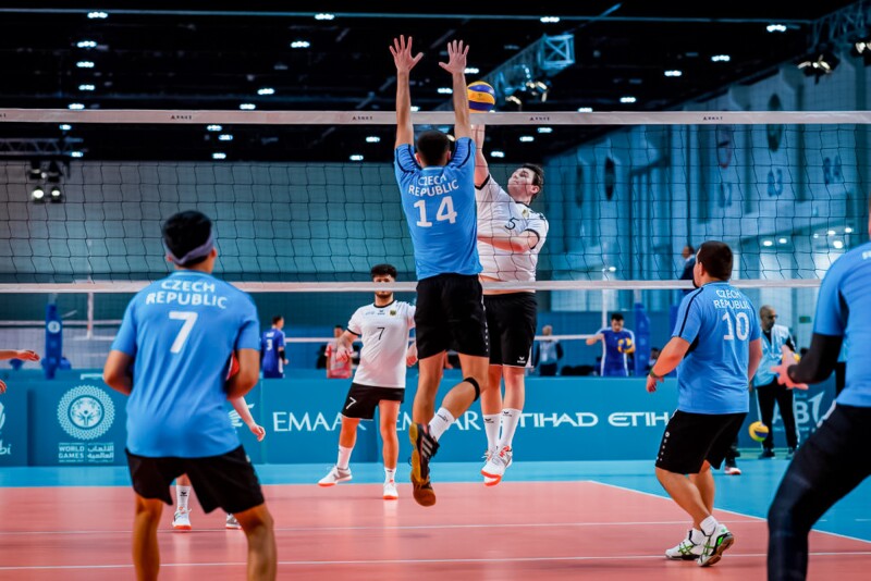 Das Foto zeigt zwei Mannschaften beim Volleyball. Zwei Spieler sind in Aktion am Netz. Ein Spieler versucht den Ball über das Netz zu spielen, sein Gegner hat beide Arme nach oben gestreckt, um den Ball abzuwehren.