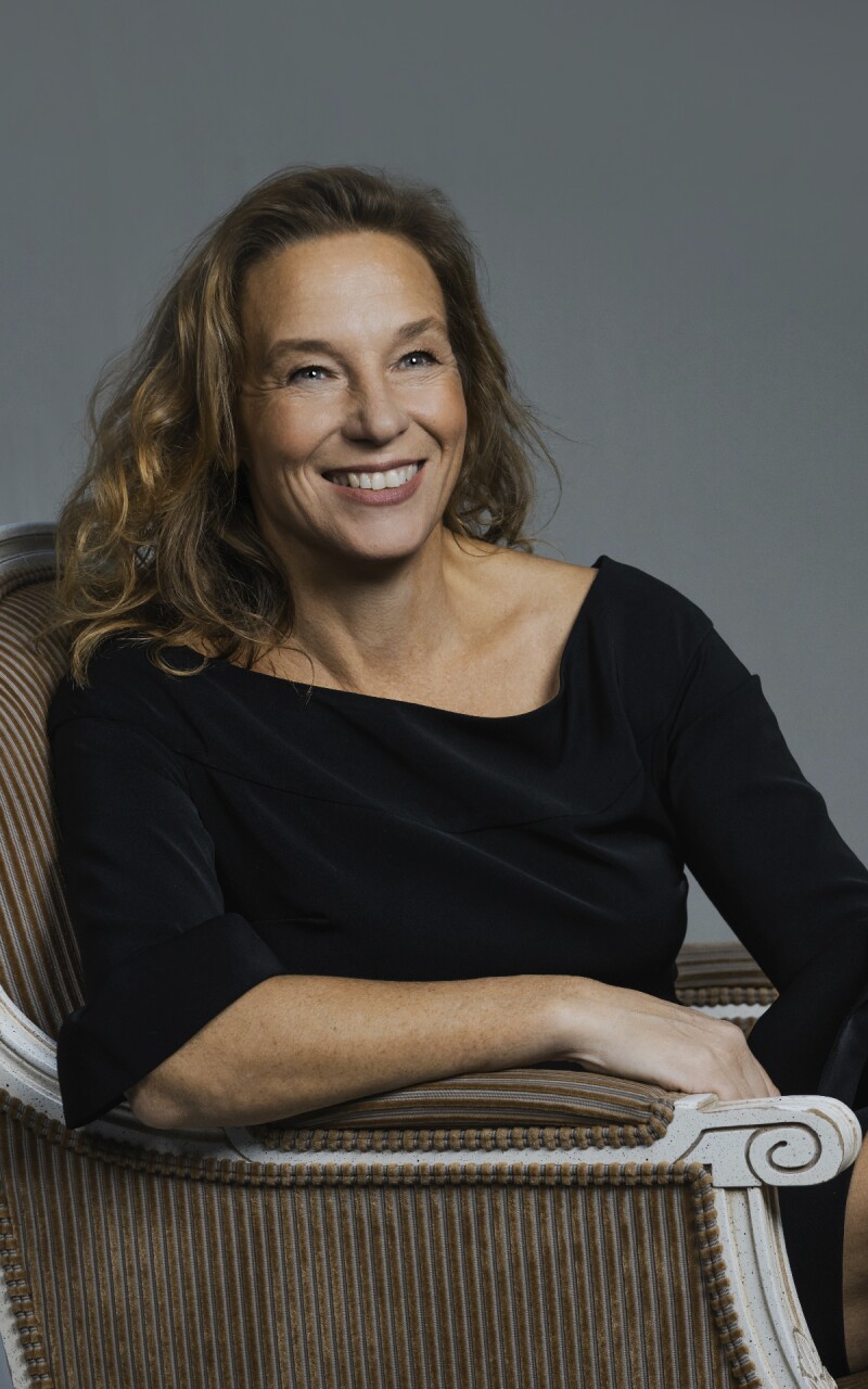 Auf dem Foto ist die deutsche Modedesignerin Anja Gockel zu sehen. Sie sitzt in einem Sessel, trägt ein schwarzes Kleid und lacht vergnügt. 