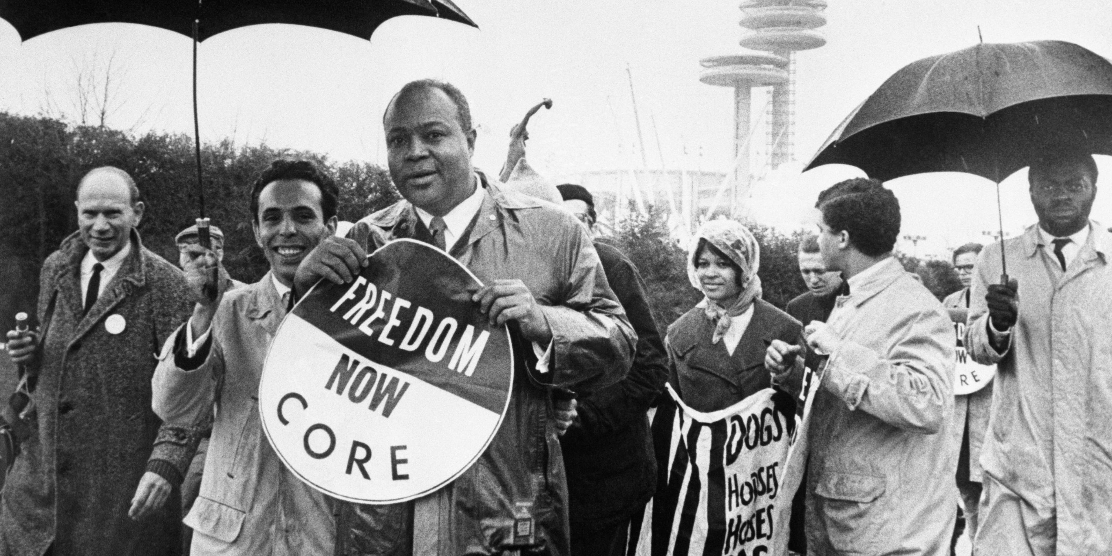 James Farmer, national director of the Congress of Racial Equality, holds a large sign as he leads a CORE demonstration during opening day ceremonies at the World's Fair. In the background is the New York State Pavilion.