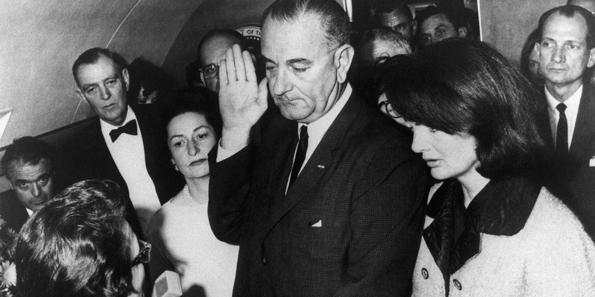 Vice President Lyndon B. Johnson is sworn in to the office of the Presidency aboard Air Force One in Dallas, Texas, hours after the assassination of President John F. Kennedy. Johnson is flanked by wife, Lady Bird Johnson (L), and First Lady Jacqueline Kennedy during the ceremony, which is being administered by U.S. District Judge Sarah Hughes. At farthest left in the background is Jack Valenti.