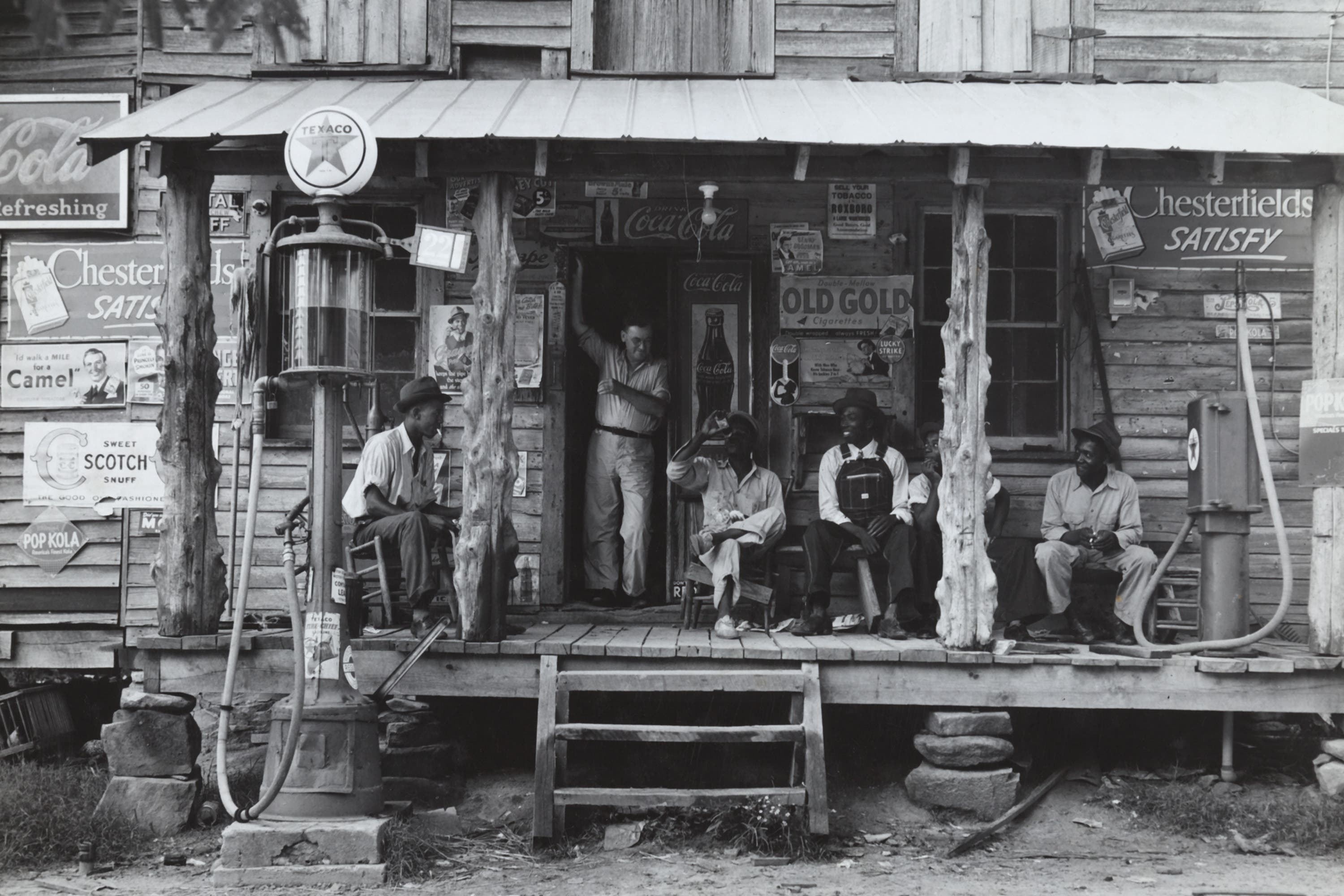 UNITED STATES - CIRCA 1939: Country store on dirt road. Sunday afternoon. Note the kerosene pump on the right and the gasoline pump on the left. Rough, unfinished timber posts have been used as supports for porch roof. Negro men are sitting on the porch. Brother of store owner stands in doorway. Gordonton, North Carolina (Photo by Buyenlarge/Getty Images)