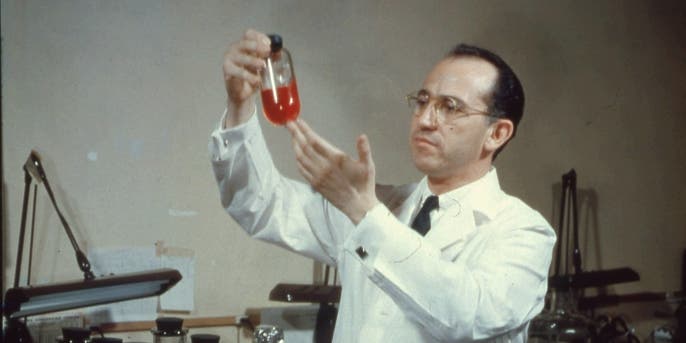 View of American scientist and physician Jonas Salk (1914 - 1995), developer of the polio vaccine, wearing a white lab coat, and smiling while holding up a bottle in the laboratory, mid twentieth century. (Photo by PhotoQuest/Getty Images)