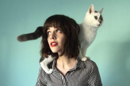 Poet Paige Lewis with short brown hair in a button down shirt and a white cat on their shoulders