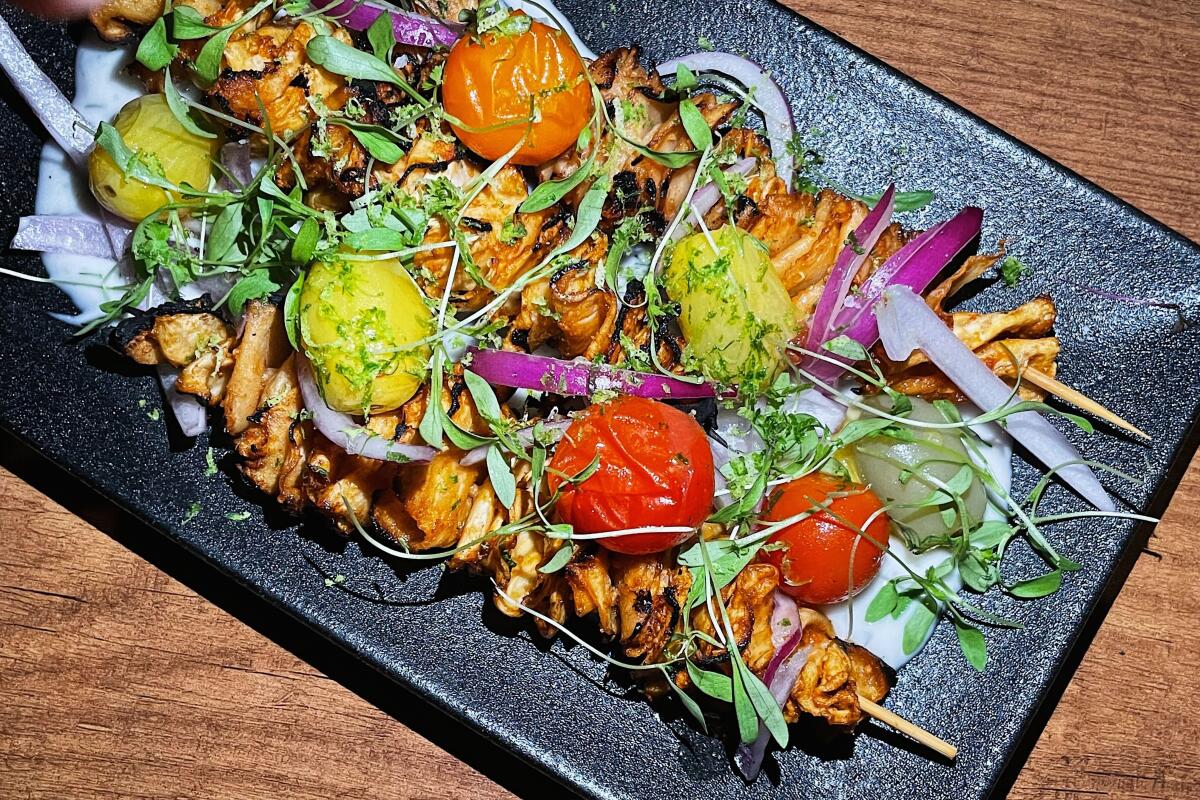 Two celery root suya skewers on a black plate, topped with tomatoes and herbs