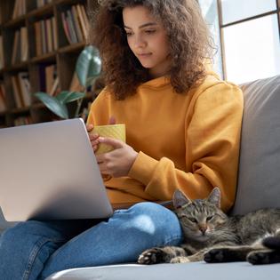 A young woman sits on a sofa with her pet cat and watches a TV show on her laptop. VPNs can help you unblock foreign streaming content.