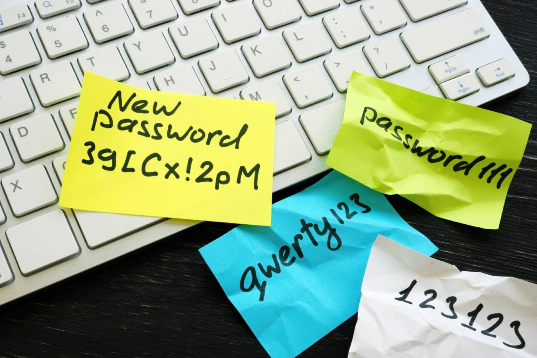 Near a white computer keyboard are several weak passwords written on sticky notes plus one with a new strong password.