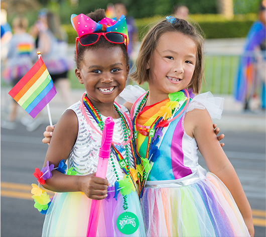 2 little girls in rainbow dresses with rainbow flags smiling at the camera.