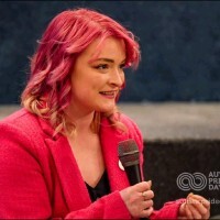 A woman with pink hair speaking into a microphone. She is wearing a red jacket and a pin on her lapel, and she appears to be engaged in a conversation or public speaking event