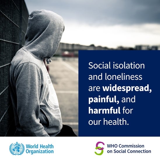 An unidentifiable person, wearing a hoodie with the hood covering their head and face, leaning against a fence looking away from the camera. Caption: 'Social isolation and loneliness are widespread, painful, and harmful for our health'.